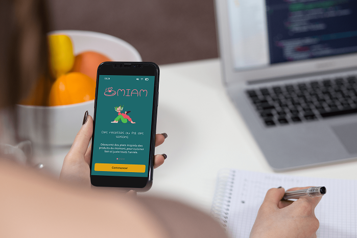 Mockup de l’application culinaire Miam affiché sur un smartphone tenu en main, montrant l’écran d’accueil avec le logo, l’illustration et le bouton « Commencer ».